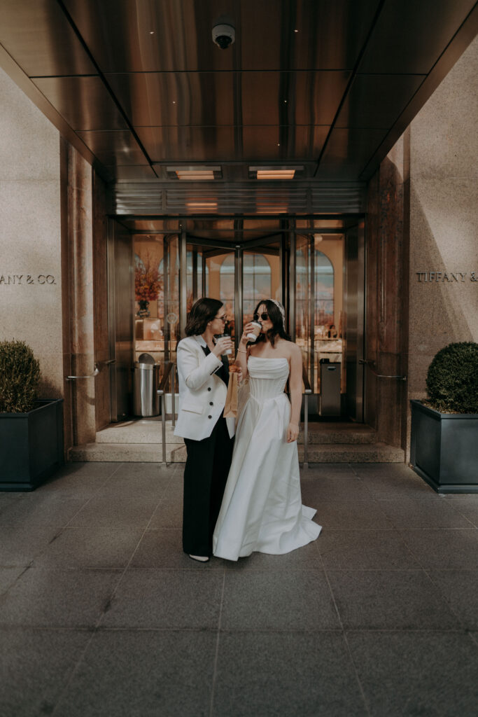 Two brides in front of Tiffany and Co on Fifth Avenue NYC during intimate elopement morning session, holding coffee and pastries in the early morning light