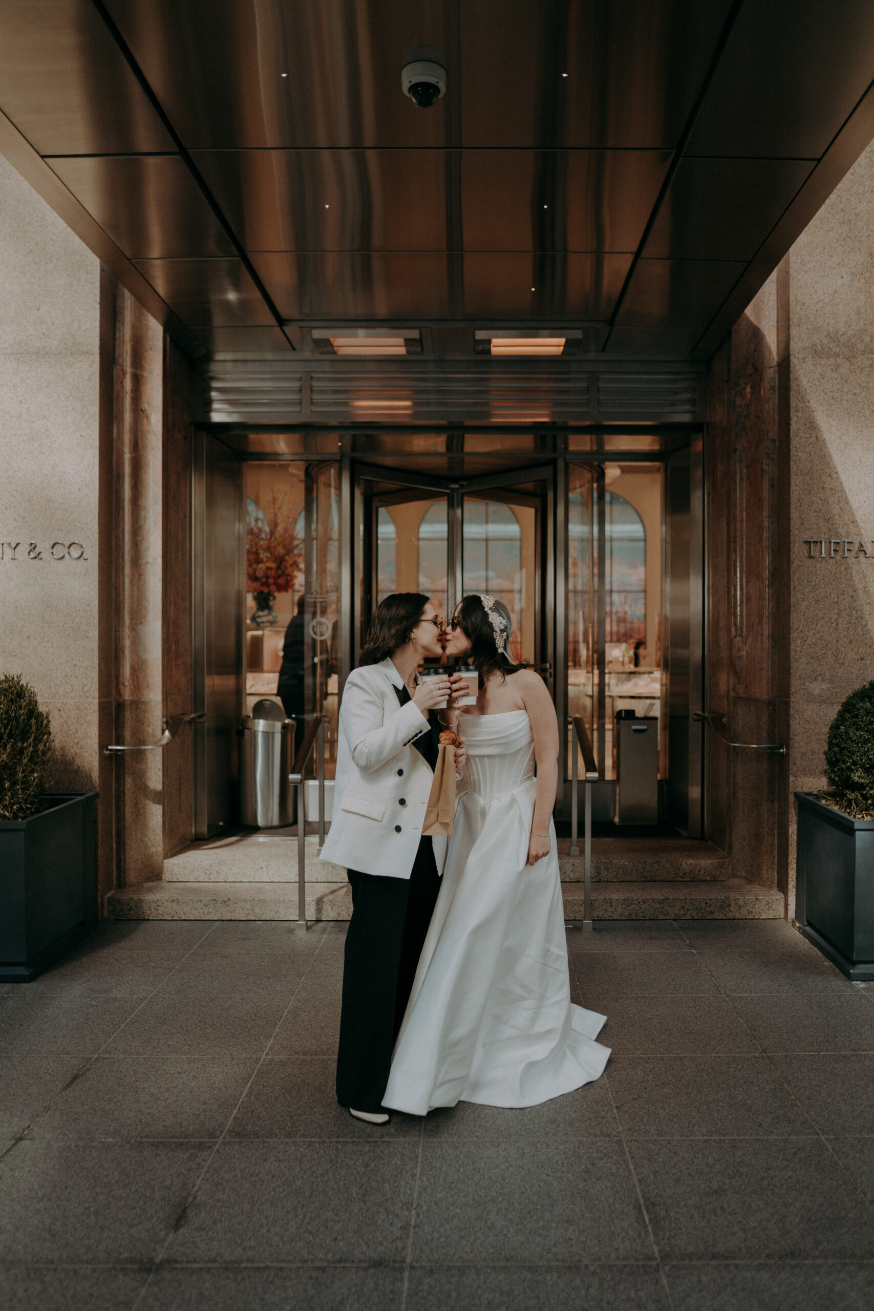 Two brides in front of Tiffany and Co on Fifth Avenue NYC during intimate elopement morning session, holding coffee and pastries in the early morning light