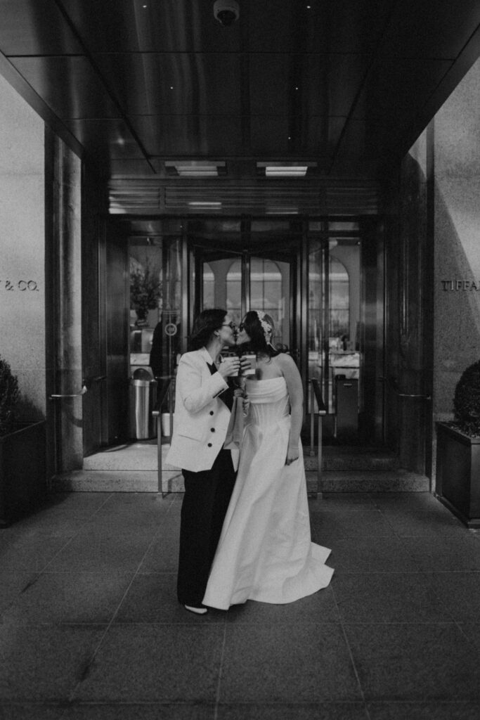 Two brides in front of Tiffany and Co on Fifth Avenue NYC during intimate elopement morning session, holding coffee and pastries in the early morning light