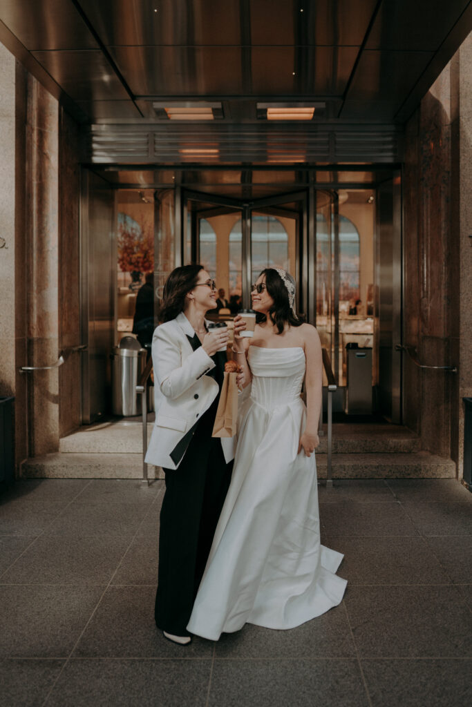 Two brides in front of Tiffany and Co on Fifth Avenue NYC during intimate elopement morning session, holding coffee and pastries in the early morning light
