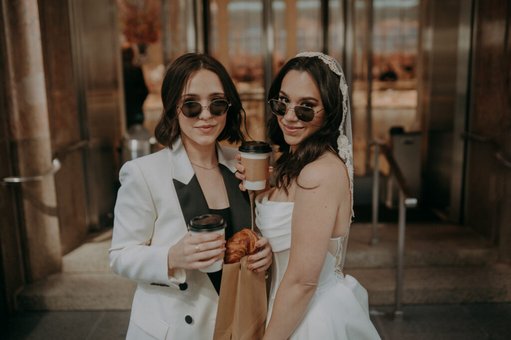 Two brides in front of Tiffany and Co on Fifth Avenue NYC during intimate elopement morning session, holding coffee and pastries in the early morning light