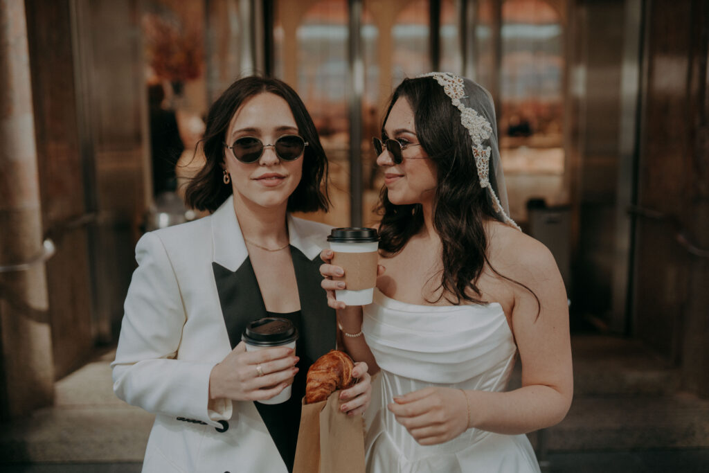 Two brides in front of Tiffany and Co on Fifth Avenue NYC during intimate elopement morning session, holding coffee and pastries in the early morning light