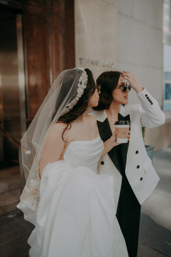 Two brides in front of Tiffany and Co on Fifth Avenue NYC during intimate elopement morning session, holding coffee and pastries in the early morning light