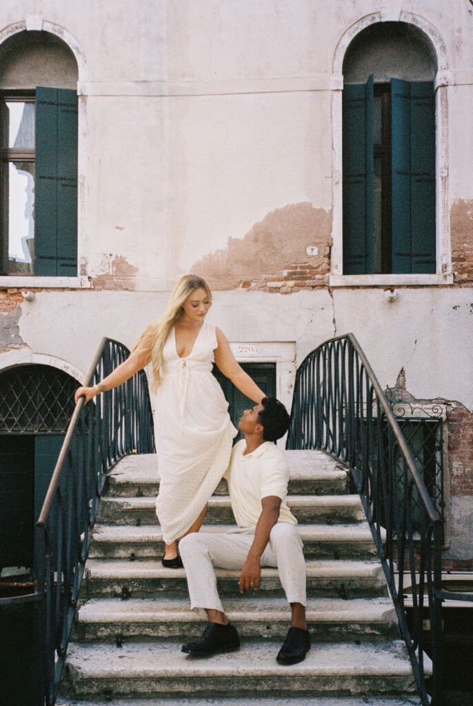 Couple on a Venice bridge staircase, her in a white dress descending, him seated looking up at her