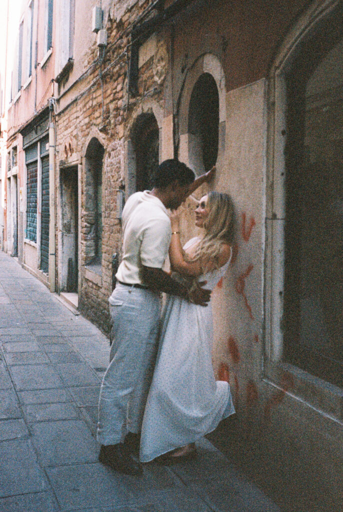 Couple embracing in a quiet Venice alley, him leaning in to kiss her against a weathered stone wall, her in a white dress