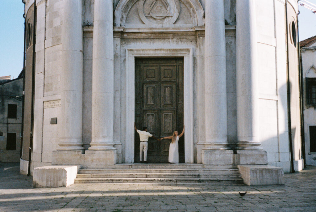 Couple standing beneath a large church doorway in Venice, hands touching in the middle